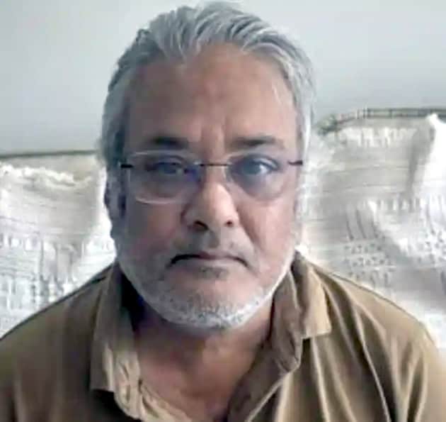 تصویر میں ایک مرد، گرے بال اور چشمہ کے ساتھ، آرام دہ لباس میں، ایک داخلی یا بیرونی جگہ میں نظر آ رہا.