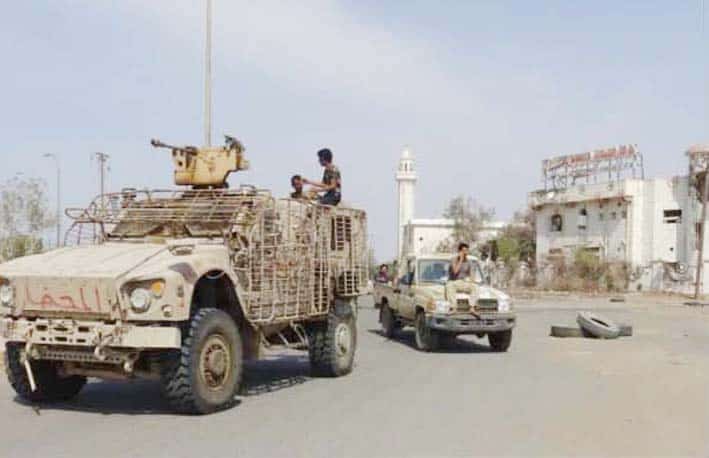 عسکری اور فوجی گاڑی ایک سڑک پر کھڑی ہیں، پس منظر میں عمارتیں اور ایک مینار ہے.