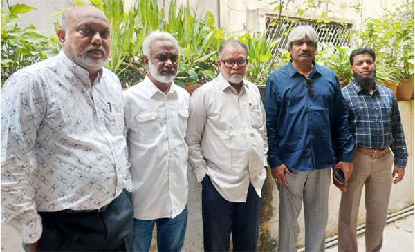 پانچ مرد کھڑے ہیں، سب کے پس منظر میں سبز پودے ہیں.